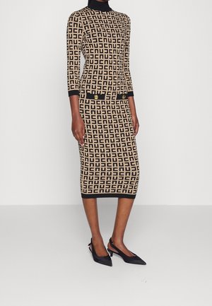 Woman wearing a beige and black patterned knee-length dress with three-quarter sleeves and black pointed slingback heels.