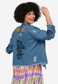 Jeansjacke mit Frontknöpfen, used Details und buntem gesticktem Text. Verfügt über schwarze Textgrafiken auf dem Rücken und den Ärmeln.