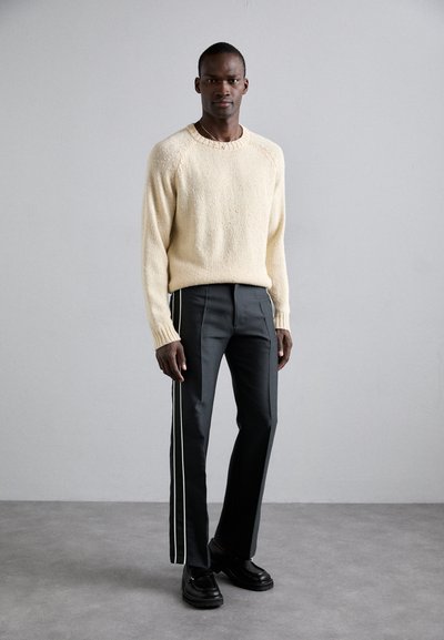 Beige gebreid sweater met textuurdetail, gecombineerd met slim-fit donkere broeken met witte zijkanten en zwarte schoenen.