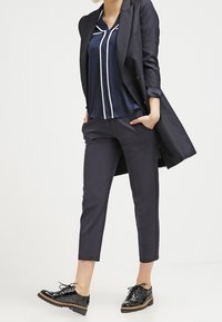 Marinblus i satin med vit kantning, skräddarsydd svart kavaj, korta svarta byxor och glänsande svarta skor med snörning.