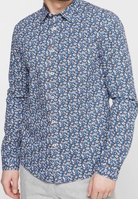 Homme portant une chemise à manches longues bleue avec un motif floral blanc et marron, boutonnée, associée à un pantalon gris clair.