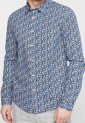 Homme portant une chemise à manches longues bleue avec un motif floral blanc et marron, boutonnée, associée à un pantalon gris clair.