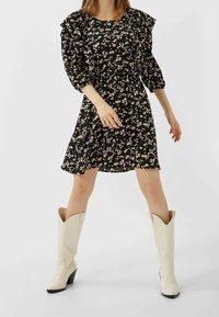 Robe noire à fleurs avec des manches à volants, taille cintrée et jupe évasée. Portée avec des bottes de cowboy blanches montant jusqu'aux genoux.