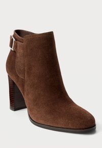 Stivale ankle in suede marrone con punta appuntita e tacco spesso. Caratterizzato da una fascia in pelle con una fibbia argentata alla caviglia.