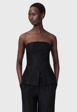 Γυναίκα με κοντά μαλλιά που φοράει ένα μαύρο τοπ χωρίς τιράντες και μαύρο παντελόνι, στέκεται με τα χέρια στις τσέπες κοιτώντας προς τα πλάγια.