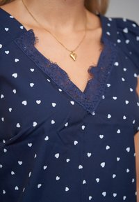 Námořnicky modrá halenka s bílým vzorem srdíček. Krajkový lem u výstřihu do V. Měkká látka, krátké balónové rukávy a volný střih. Zlatý náhrdelník ve tvaru srdce.