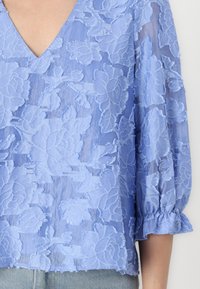 Lys blå bluse med blomster-lace tekstur, V-hals og flagrende klokkeslæt. Blødt, gennemsigtigt stof med intrikate broderidetaljer.
