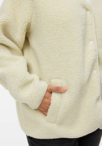 Chaqueta de forro polar color crema con una textura suave, que cuenta con un cierre de botones y bolsillos laterales. Incluye puños y dobladillo acanalados.