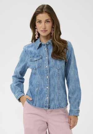 Femme portant une chemise en jean bleue boutonnée avec deux poches poitrine et un pantalon rose, accessoirisée avec de longues boucles d'oreilles roses, posant avec une main dans la poche.