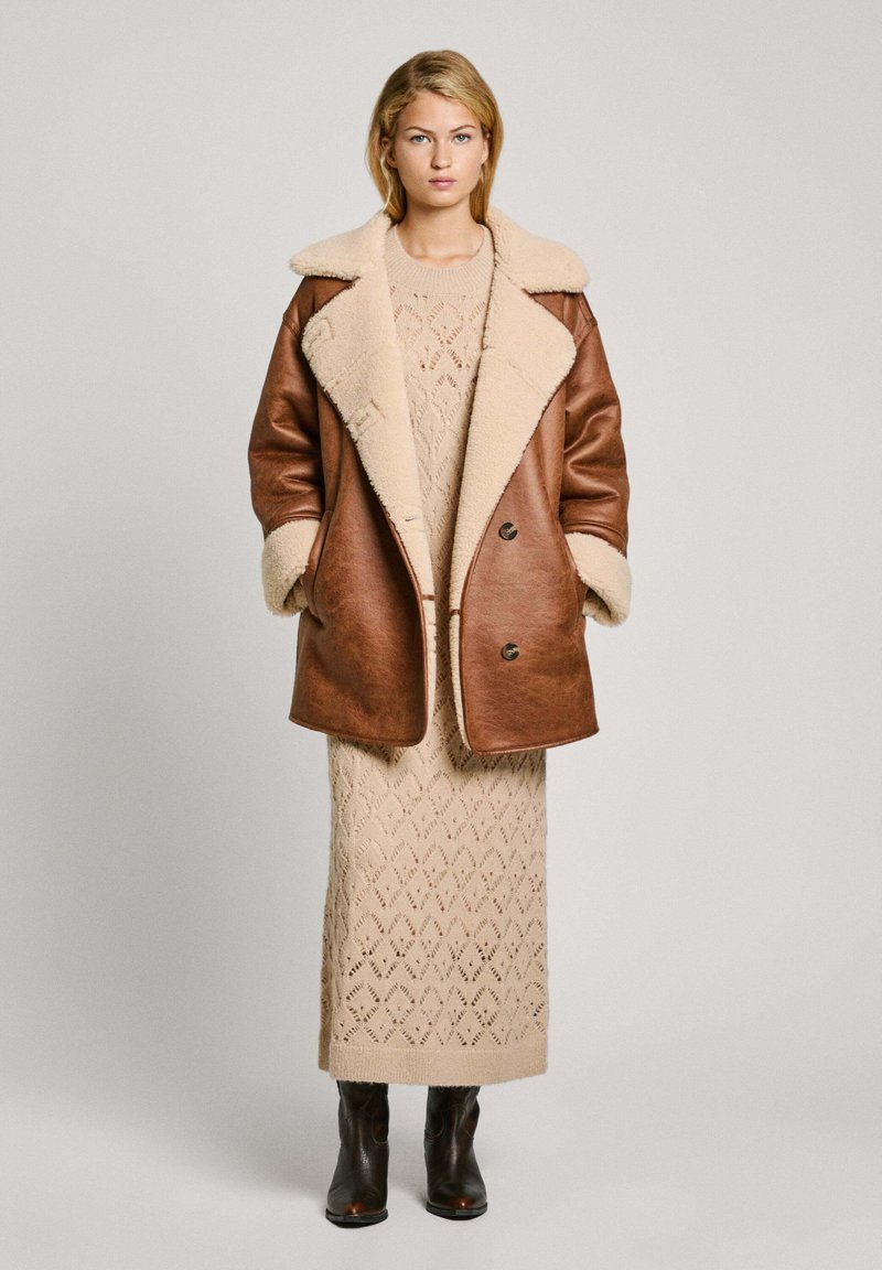 Manteau en cuir marron avec col et poignets en shearling crème, superposé sur une robe tricotée beige à motif en losange, complétée par des bottes noires.