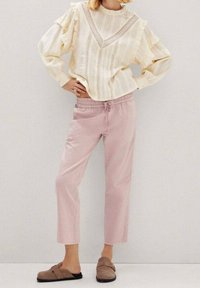 Helle, strukturierte Crèmetbluse mit langen Rüschenärmeln und einem V-förmigen Spitzen-Detail, kombiniert mit weichen, rosa Trousers mit elastischem Bund und tanfarbigen Slippern.