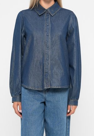 Femme portant une chemise en jean bleue à boutons, manches longues avec col et un pantalon en jean bleu assorti, debout devant un fond uni.