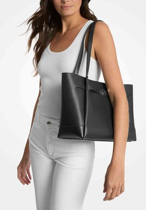 Schwarze Ledertasche mit strukturiertem Finish, flachem Boden, langen Trägern und einem schlichten Akzentdetail. Getragen zu einem weißen Tanktop und Hosen.