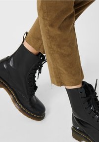 Bottines noires à lacets en cuir lisse, bout rond et surpiqûres jaunes sur la semelle, associées à un pantalon en velours côtelé marron.