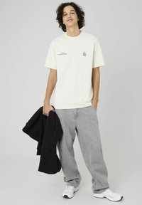 T-shirt crème avec un graphisme bleu, jeans gris ample, baskets blanches. Sweatshirt noir tenu dans la main gauche, coupe droite, ajustement décontracté.