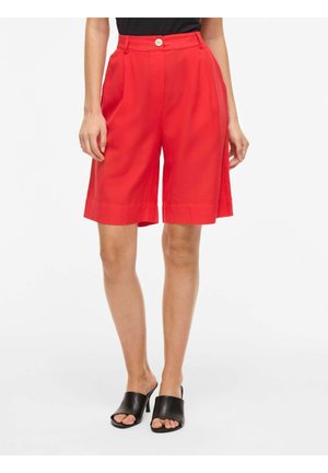 Vrouw draagt hoge taille, felrode, op maat gemaakte shorts tot op de knie met knoopsluiting en zwarte open-teen hakmuiltjes.