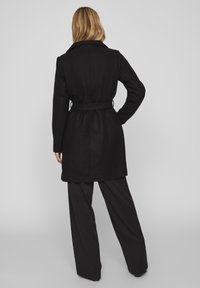 Cappotto in misto lana nero con vita cinturata, ampio colletto e cuciture verticali, abbinato a pantaloni neri a gamba larga.