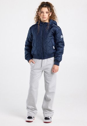 Kind met krullend haar draagt een marineblauw doorgestikt jack, lichtgrijze joggingbroek en zwart-witte sneakers, staand tegen een witte achtergrond.