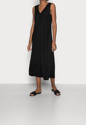 Schwarzes ärmelloses Midikleid mit V-Ausschnitt, weichem Stoff, lockerem Schnitt und subtilen Drapierungen. Kombiniert mit schwarzen flachen Sandalen auf hellem Untergrund.