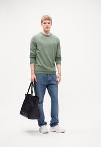 Green sweatshirt, blue jeans, white sneakers, holding black tote bag. Sweatshirt features small logo; bag has embossed text. Simple, casual attire.