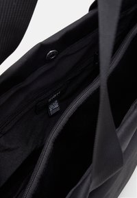 Interno di una borsa a spalla in tessuto nero che mostra scomparti con zip e un'etichetta con il marchio e codice QR all'interno.