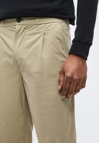 Άνδρας που φοράει μπεζ πιετέ παντελόνια και μαύρο μακρυμάνικο πουκάμισο, με έμφαση στη μέση και το δεξί χέρι που ακουμπάει δίπλα στο πόδι.