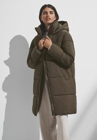 Manteau doudoune en kaki avec fermeture éclair et capuche. Présente des panneaux cousus et est de longueur genou, fabriqué à partir d'un tissu doux et mat.