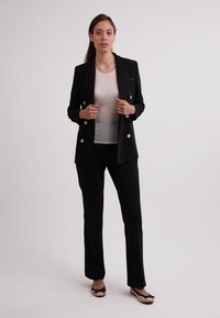 Schwarzer, taillierter Blazer mit doppelreihigem Design und silbernen Knöpfen, kombiniert mit einem weißen Oberteil und schwarzen Hosen; flache Schuhe runden das Outfit ab.