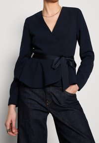 Námořnický wrap top s dlouhými rukávy, designem peplum a detaily z lesklé saténové stuhy. Stylizováno s tmavými džínovými kalhotami, které vykazují hladkou texturu.