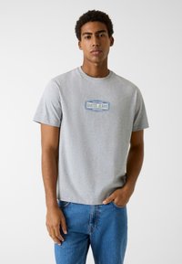 Szara bawełniana koszulka z okrągłym dekoltem, z niebiesk�ą naszywką z logo "GUESS JEANS" na przodzie. Noszona z niebieskimi dżinsami.