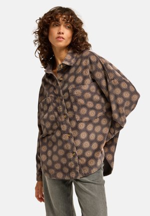 Vrouw met krullend haar, die een losse, bruine blouse met knopen en een zonpatroon draagt en een grijze broek, poserend met één hand achter haar rug.