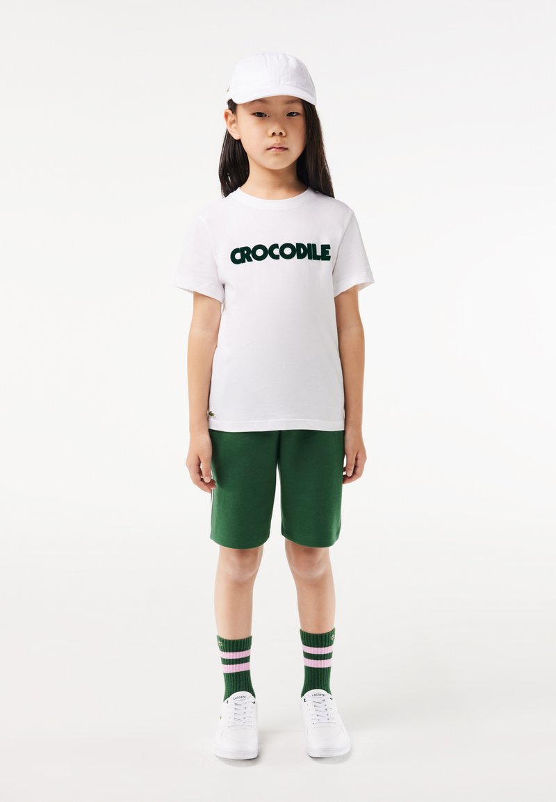 T-shirt en coton blanc avec le texte "CROCODILE" en vert, short vert, baskets blanches et chaussettes vertes avec des rayures roses. Casquette blanche.