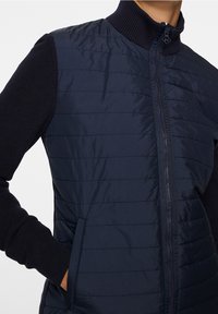 Marinblå quiltad väst med hög krage, ribbstickade ärmar och sidofickor. Har horisontell sömnad för textur och en dragkedja fram.
