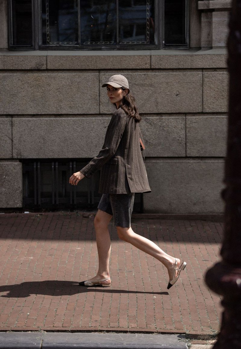 Bruine pinstriped blazer, lichtgrijze pet, denim shorts en twee-kleurige slingback schoenen. Model loopt op een stenen pad nabij een muur van steen.