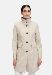 Beiger Trenchcoat mit hohem Kragen, Frontknöpfen und zwei Seitentaschen. Glattes Material und taillierte Passform.