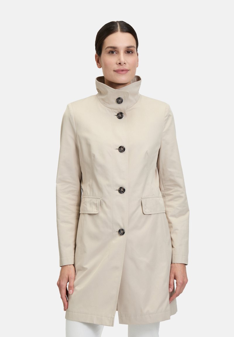 Beiger Trenchcoat mit hohem Kragen, Frontknöpfen und zwei Seitentaschen. Glattes Material und taillierte Passform.