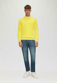 Gelber Pullover mit rundem Ausschnitt, kombiniert mit blauen Jeans und weißen Turnschuhen. Der Stoff wirkt weich und hat eine einfarbige Optik.