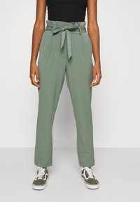 Pantalons vert sur mesure avec taille à nouer, présentant une texture lisse, une coupe droite et des poches latérales, associés à des baskets blanches et vertes.