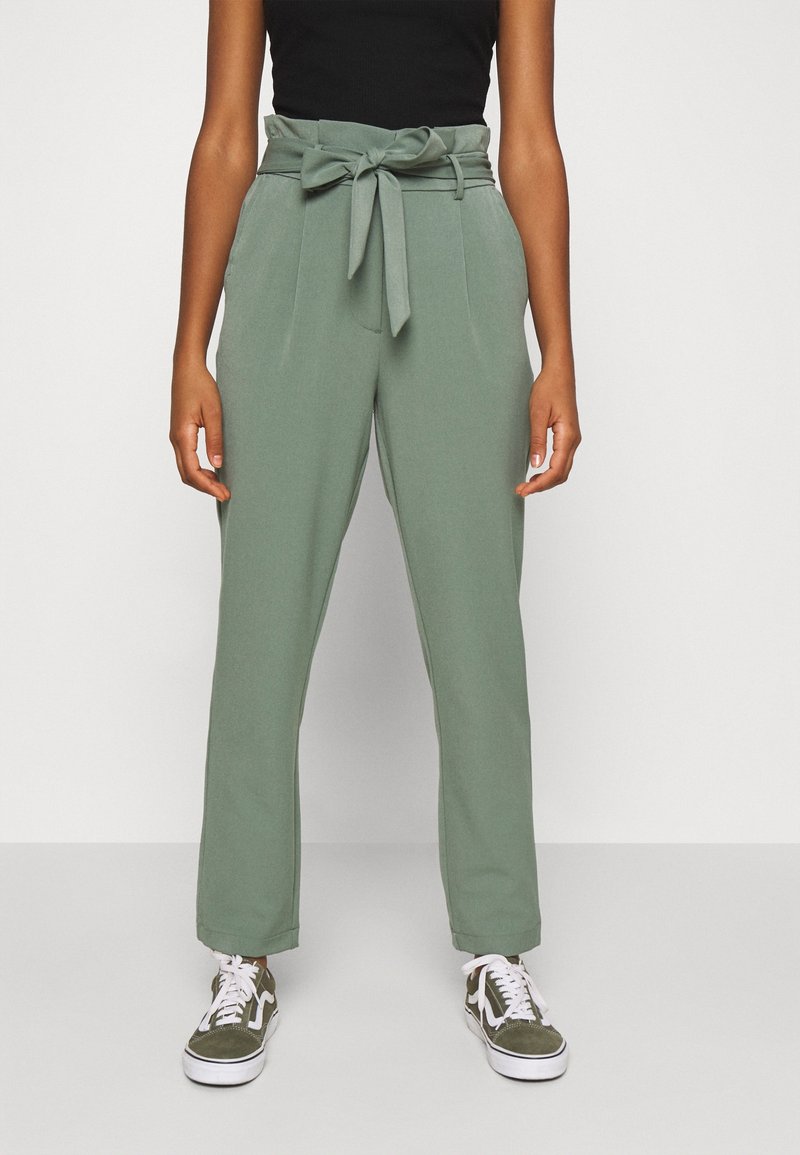 Pantalons vert sur mesure avec taille à nouer, présentant une texture lisse, une coupe droite et des poches latérales, associés à des baskets blanches et vertes.