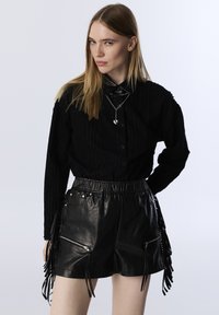 Zwart gestructureerd overhemd met knopen, gecombineerd met zwarte leren shorts met een hoge taille, voorzien van franjes en ritszakken. Zilveren sieraden inbegrepen.