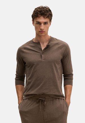 Jonge man met krullend haar die een bruin Henley-shirt met lange mouwen draagt en bijpassende broek met trekkoord, handen in de zakken.