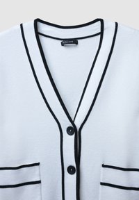 Cardigan bianco con accenti neri, caratterizzato da un collo a V, due tasche frontali e chiusura a bottoni. Texture morbida con dettagli di rifinitura a contrasto.