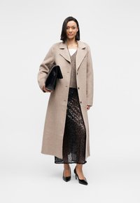 Μπεζ μάλλινο παλτό με γιακά με σχισμή, μεγάλα κουμπιά και πλαϊνές τσέπες· φορεμένο πάνω από καφέ πουλόβερ και μαύρη φούστα με παγιέτες. Μαύρο τσαντάκι και ψηλοτάκουνα παπούτσια.