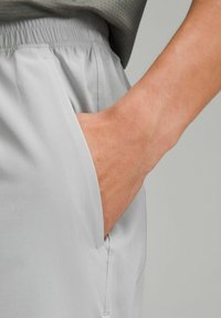 Lysegrå sportshorts med elastisk talje og sidelommer, vist med en hånd hvile i den højre lomme for at give en fornemmelse af størrelsen.