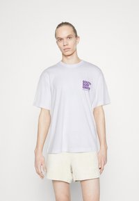 Balta medvilnė marškinėliai trumpomis rankovėmis ir apvalia apykakle. Kairėje krūtinės pusėje pavaizduotas violetinis spausdintas logotipas. Modelis dėvi smėlio spalvos šortus.