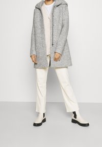 Grå kappa i ullblandning med huva och knäppt fram, kombinerad med en beige stickad tröja och off-white byxor. Svarta ankelboots med klack fullbordar looken.