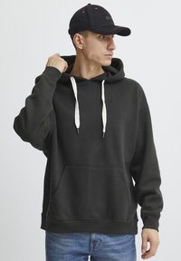 Felpa nera con tasca frontale, cordini bianchi e polsini a coste. Abbinata a jeans blu e un cappellino nero.