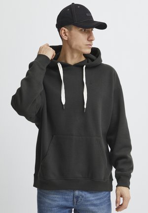 Svart hettegenser med frontlomme, hvite snorer og ribbestrikkede mansjetter. Kombinert med blå jeans og en svart caps.