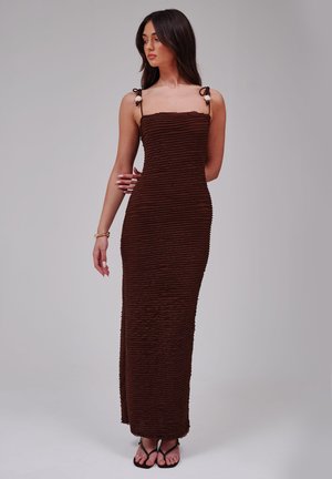 Femme portant une robe maxi marron foncé ajustée et texturée avec de fines bretelles, debout devant un fond gris neutre.