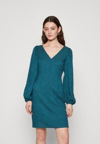 Robe teal avec un décolleté en V, des manches bouffantes longues et un tissu texturé. Coupe ajustée avec un ourlet au niveau des genoux et un détail de motif subtil.
